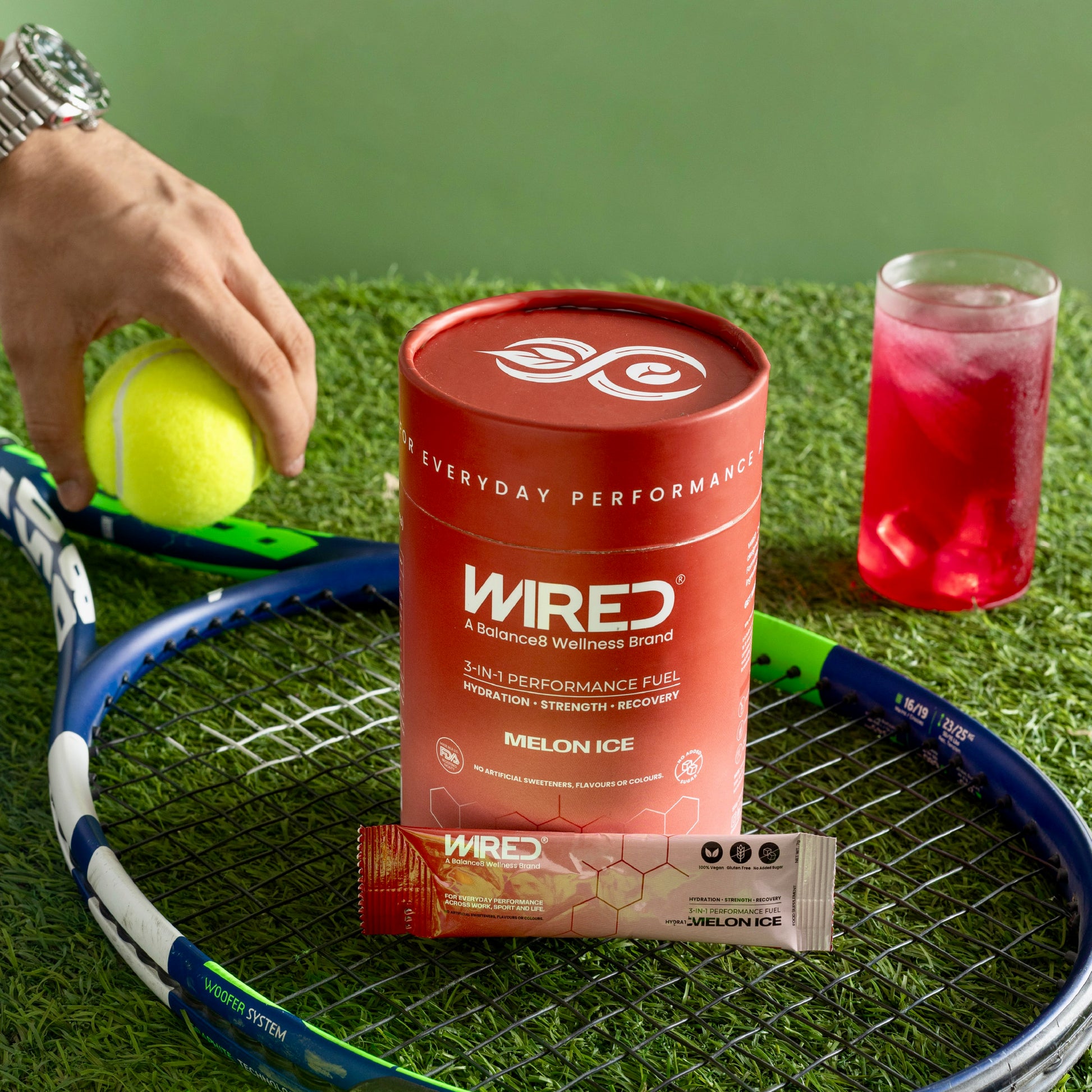 WIRED Melon Ice – sport lifestyle shot with red tub, sachet, mixed drink and tennis racket on grass | Balance8 Wellness